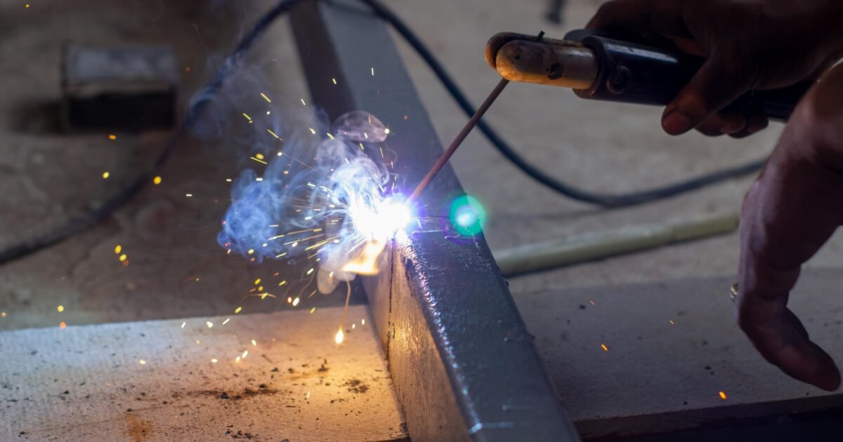 Vue rapprochée d'une personne soudant une poutre en métal, avec des étincelles lumineuses et de la fumée bleue s'élevant du point de soudure.
