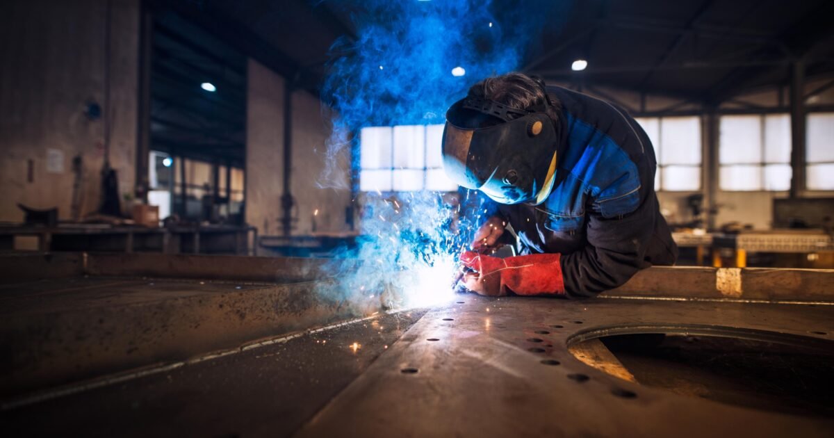 L'image montre un soudeur professionnel en uniforme de protection et casque, réalisant le soudage d'une pièce métallique dans un atelier.