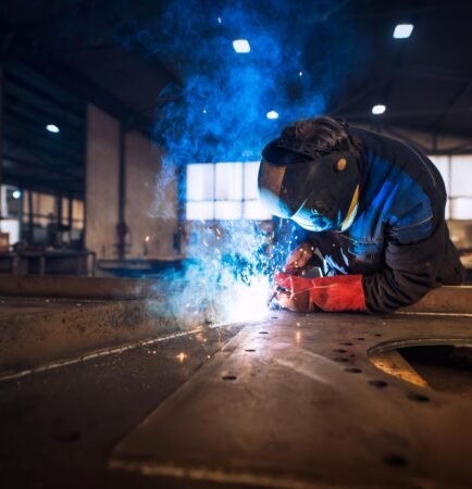 L'image montre un soudeur professionnel en uniforme de protection et casque, réalisant le soudage d'une pièce métallique dans un atelier.