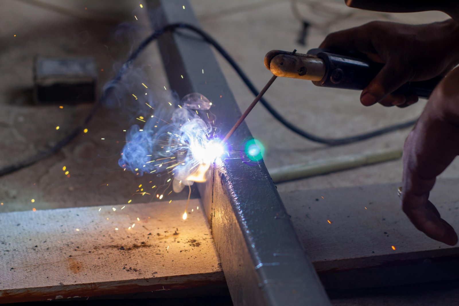 Vue rapprochée d'une personne soudant une poutre en métal, avec des étincelles lumineuses et de la fumée bleue s'élevant du point de soudure.