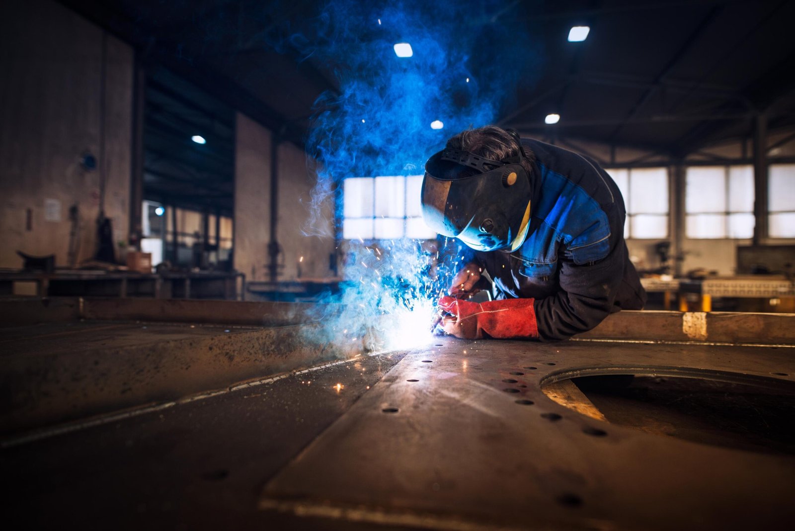 L'image montre un soudeur professionnel en uniforme de protection et casque, réalisant le soudage d'une pièce métallique dans un atelier.